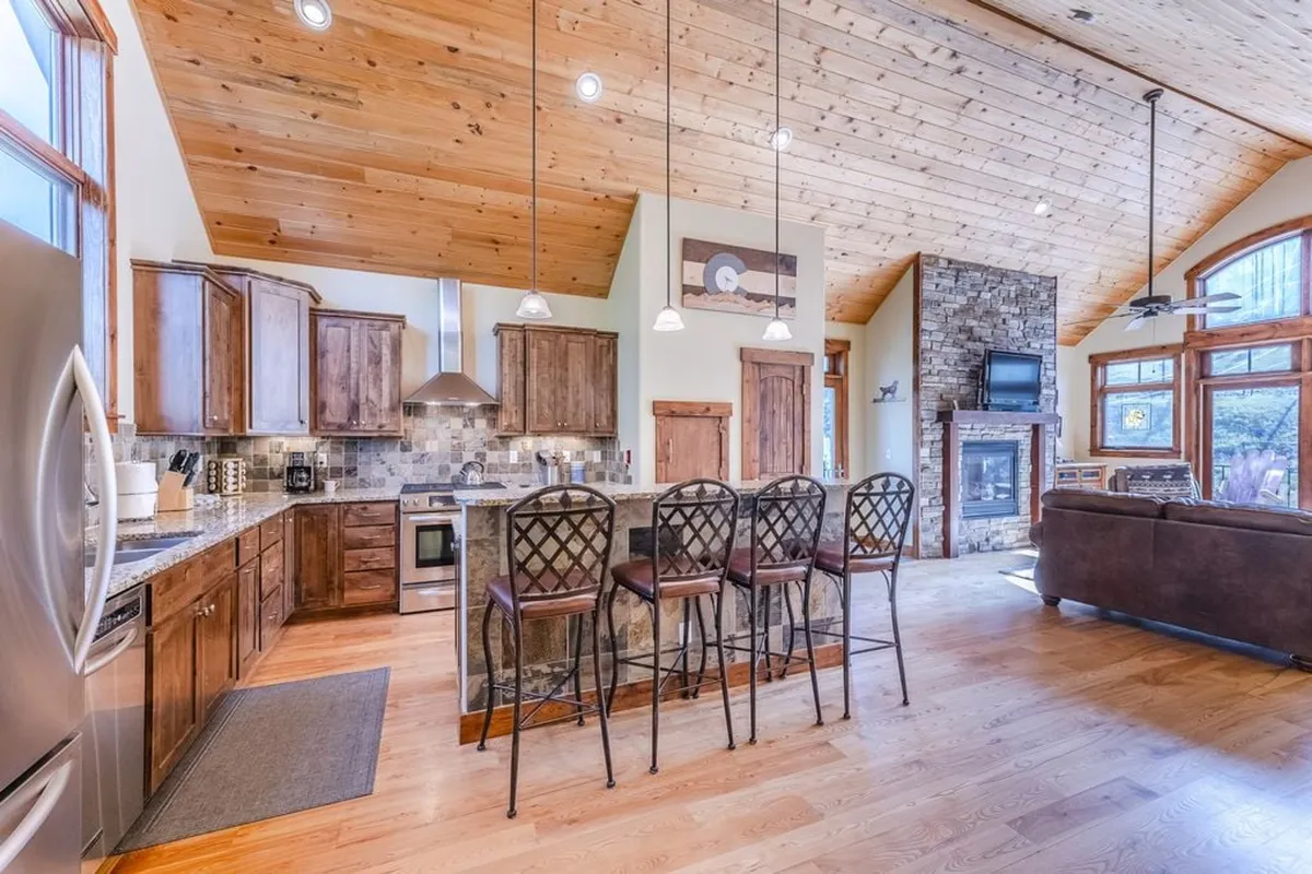 A spacious, open-concept kitchen and living area with a large kitchen island, stone fireplace, wooden cabinetry, and an inviting open layout. The scene suggests a place where cooking, entertaining, and relaxing merge, with plenty of room for group activities and celebrations.