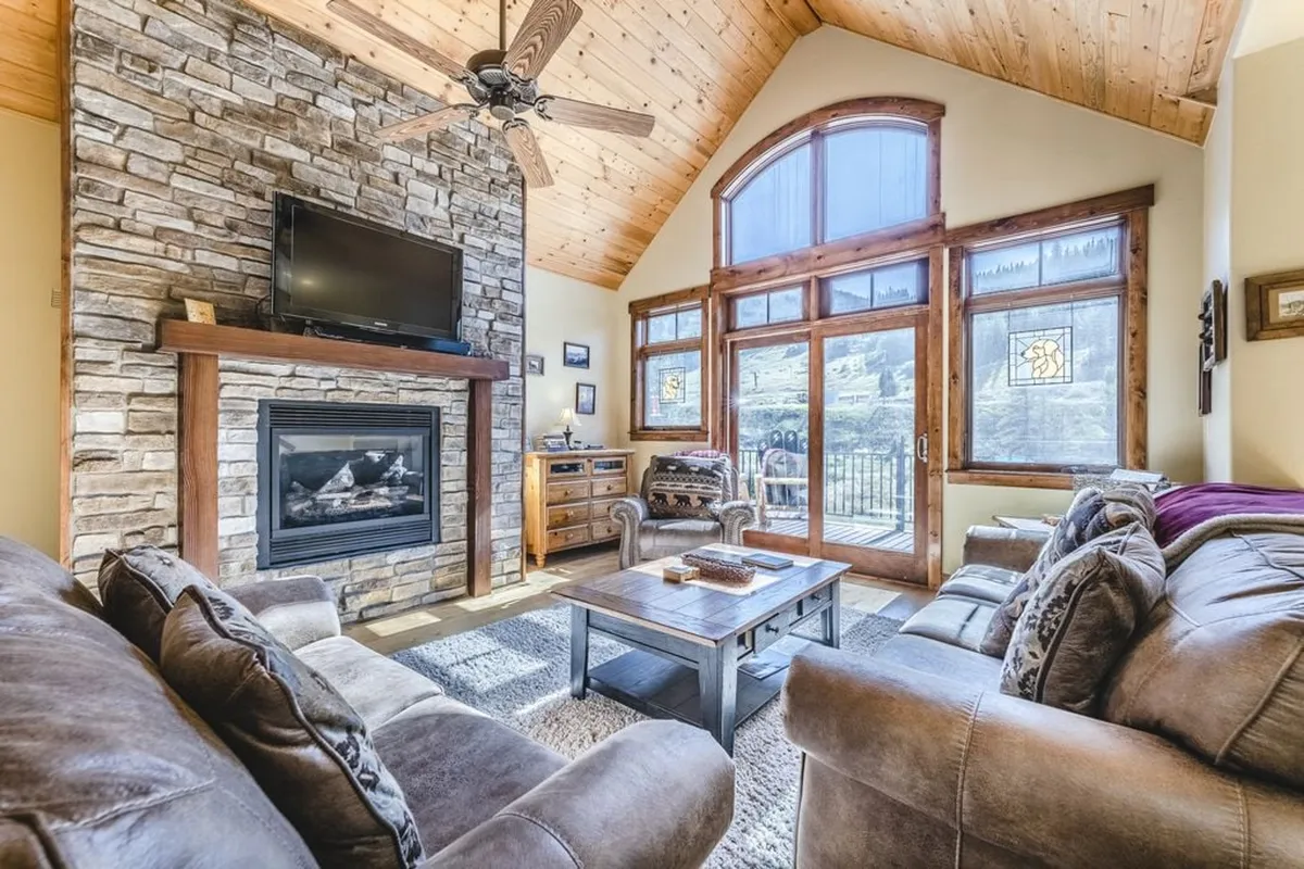 The image showcases a warm, inviting living room with a stunning stone fireplace as the centerpiece, flanked by expansive windows that reveal mountain views. Plush, comfortable seating surrounds a wooden coffee table, creating a perfect gathering spot for families or friends to relax, share stories, or enjoy the fire after a day on the slopes. The space is filled with natural light, blending cozy winter charm with the vibrant atmosphere of a mountain retreat.