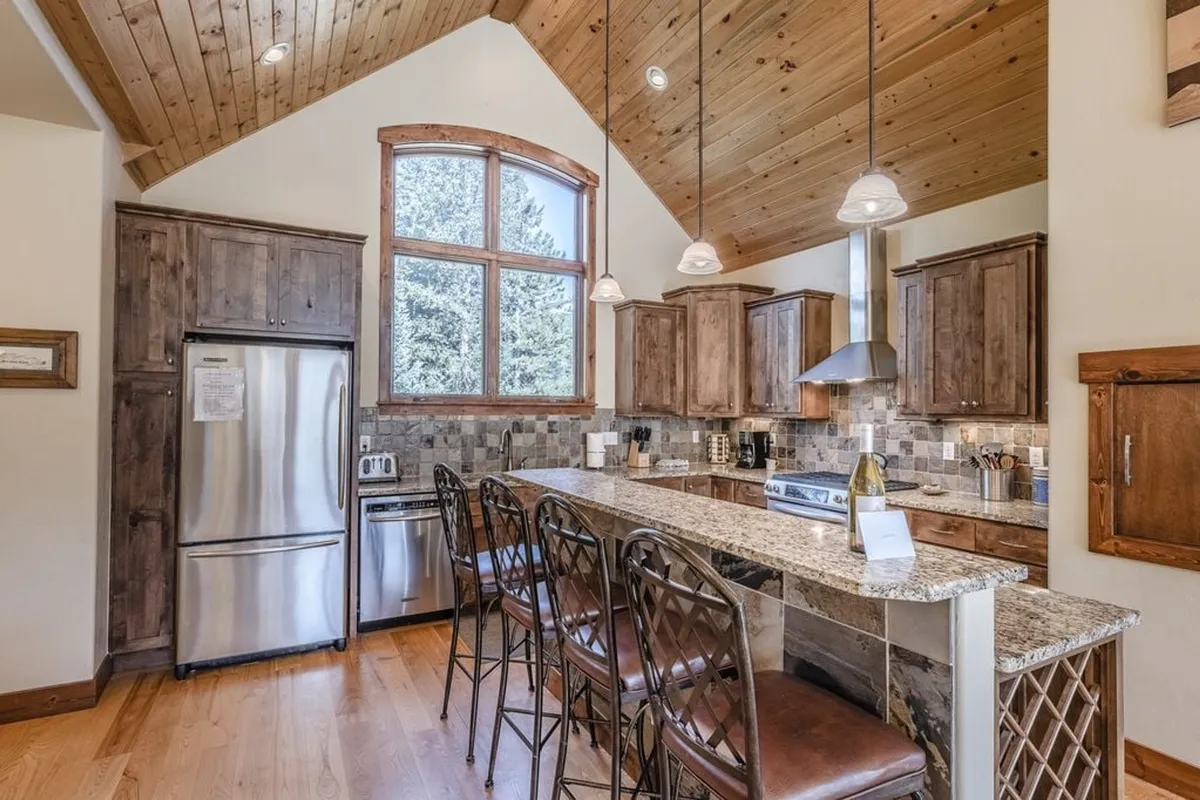 A cozy kitchen featuring a granite countertop, warm wooden cabinets, a large window allowing natural light to flood in, and bar stools lining the counter. The space is clearly designed for both cooking and gathering, with an inviting atmosphere perfect for family or group meal prep.
