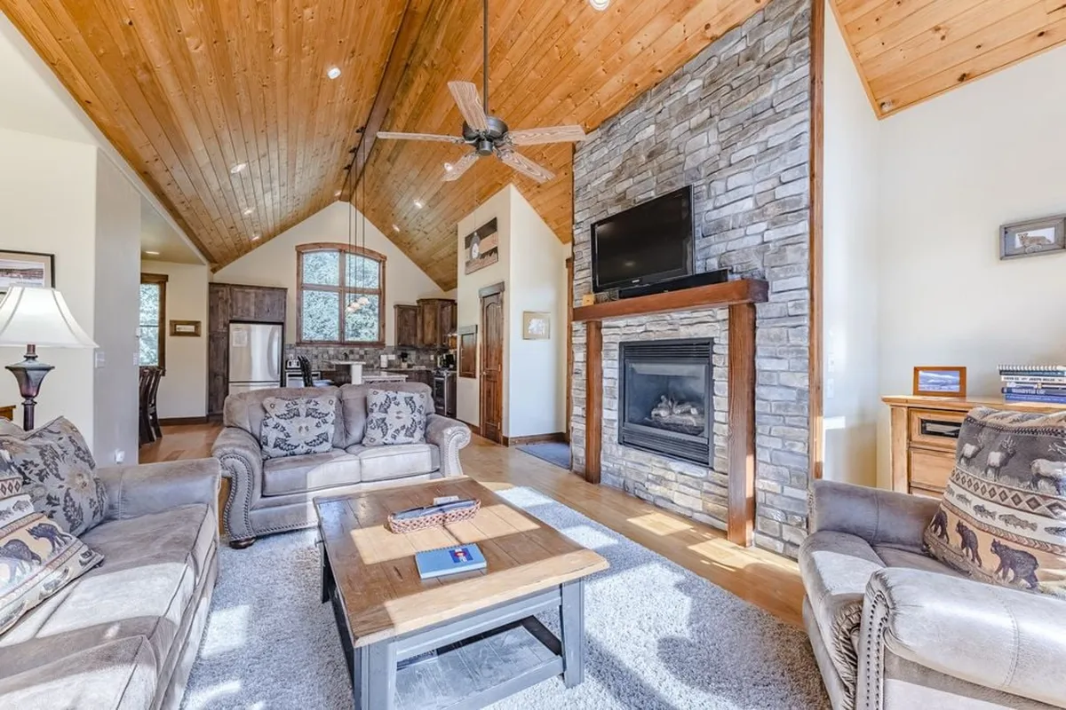 A warm, inviting living space with a stone fireplace, wood-paneled ceiling, cozy seating, and a central coffee table. The arrangement is ideal for socializing, relaxing, or having a game night by the fire.