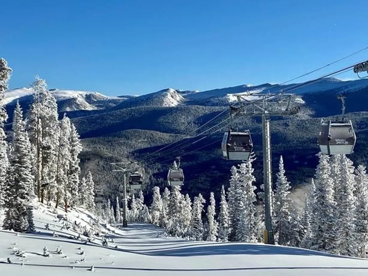 A gondola lift ascends above snow-covered mountains and pine trees as the first light of sunrise bathes the slopes, with skiers and snowboarders visible in the distance. The panorama captures both the serene beauty and the adrenaline of a day on the slopes.