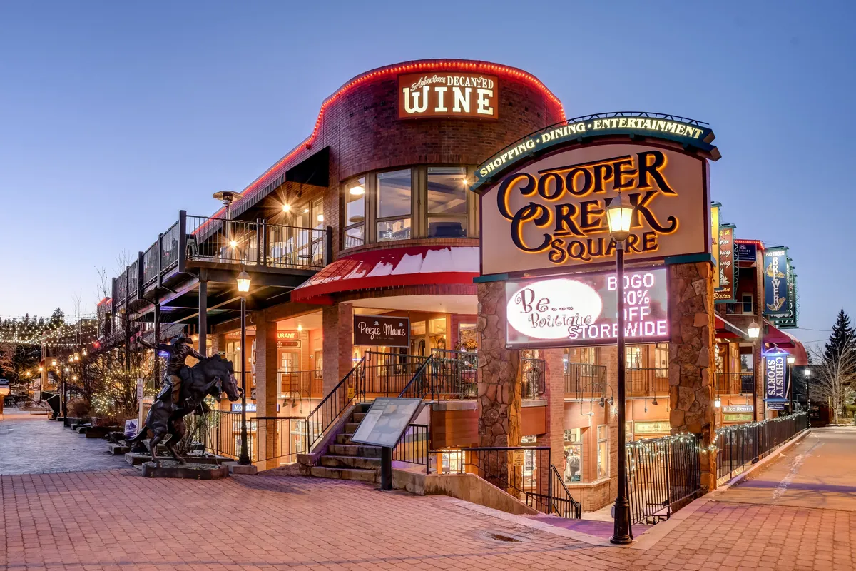 This exterior image features the iconic Cooper Creek Square sign, a wine shop, and decorative lights that create a festive ambiance. The scene captures visitors enjoying shopping, dining, and entertainment in a lively outdoor plaza, representative of Winter Park’s vibrant social and commercial life.