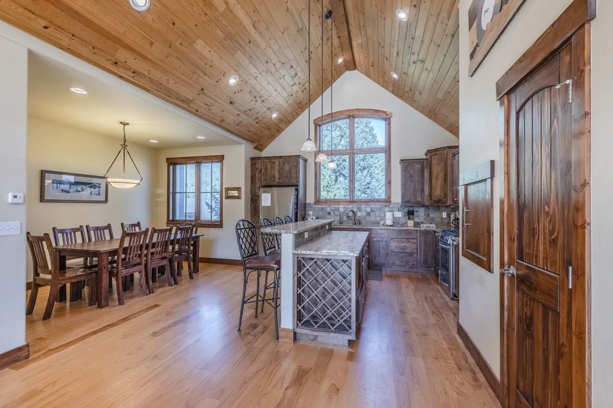 A cozy, communal dining and kitchen area inside the villa, highlighted by a large wooden dining table, island counter, and plenty of natural light from expansive windows. The space is set for cooking, dining, and playing games—perfect for group activities.