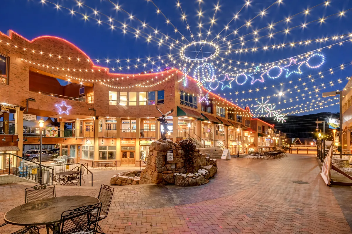A charming outdoor plaza lit by string lights and decorated with stars, featuring building facades and a stone fountain. People are gathered for outdoor dining, shopping, and socializing, evoking a joyful, festive atmosphere.