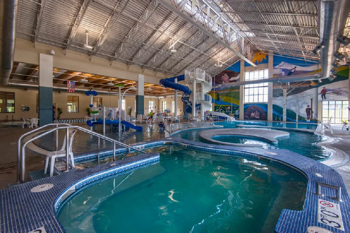 A vibrant indoor pool area at the Grand Park Recreation Center, featuring a colorful water slide, swimming pools, and children actively playing and splashing. The space is bright, energetic, and clearly family-oriented, with a playful mural on the wall enhancing the joyful atmosphere.