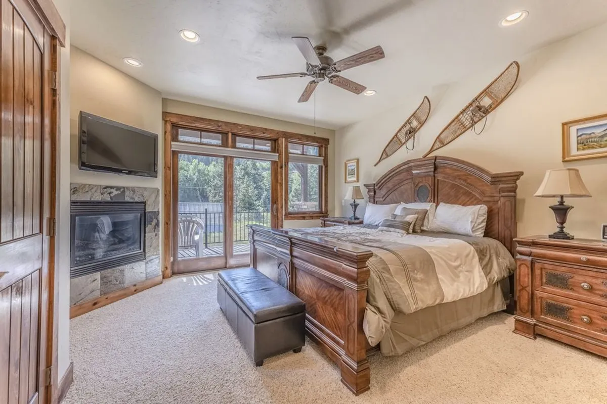 A spacious master bedroom featuring a king-sized bed with plush bedding and decorative pillows. The room includes a cozy fireplace, large windows with mountain views, and warm wooden furniture, creating an inviting retreat. Decorative snowshoes add a mountain-lodge touch, and the natural light enhances the tranquil atmosphere.
